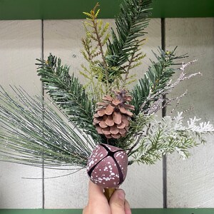May include: A festive winter bouquet with a brown pine cone, green pine branches, white frosted branches, and a brown and white bell.