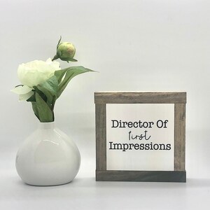 May include: A white ceramic vase holds a white peony flower and a bud with green leaves. To the right is a wooden framed sign with the text "Director Of first Impressions" in black.