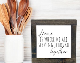 May include: A framed sign with a white background and black text that reads "Home is where we are serving Jehovah together". The sign is in a dark wood frame and is sitting on a white wooden surface.
