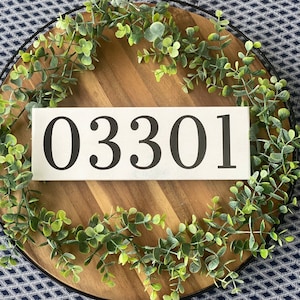 May include: A rectangular white wooden sign with the numbers "03301" in black, surrounded by a green eucalyptus garland. The sign is placed on a wooden tray with a black metal rim, set against a blue and white patterned background.