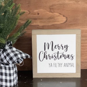 May include: A white wooden sign with a black inscription that reads "Merry Christmas Ya Filthy Animal". The sign is framed with a light brown wood frame.