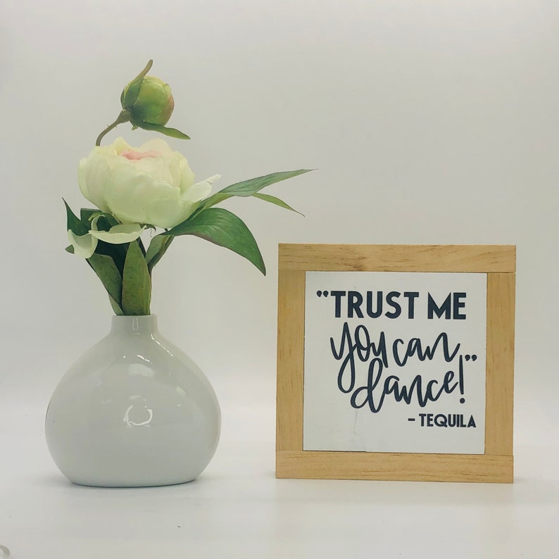 Puede incluir: Un letrero de madera con el texto "TRUST ME you can... dance! - TEQUILA" en letras negras sobre fondo blanco. El letrero est&aacute; enmarcado en madera natural y est&aacute; sobre una superficie blanca junto a un jarr&oacute;n blanco con una sola flor blanca.