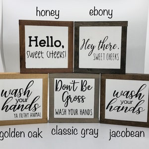 May include: Four wooden signs with different sayings and wood stains. The signs read "Hello, Sweet Cheeks!" in honey stain, "Hey there, Sweet Cheeks" in ebony stain, "Wash your hands, Ya Filthy Animal" in golden oak stain, and "Don't Be Gross, Wash Your Hands" in classic grey stain. The signs are all the same size and shape.