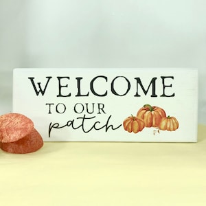 May include: White wooden sign with black text that reads "Welcome to our patch" with three orange pumpkins painted on the right side.
