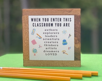 May include: A wooden sign with the text "When you enter this classroom you are" followed by a list of words including "authors", "explorers", "leaders", "scientists", "creators", "thinkers", "artists", "dreamers", and "loved". The sign is decorated with colorful illustrations of school supplies.