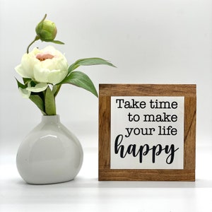 Puede incluir: Un letrero de madera con fondo blanco y texto negro que dice "T&oacute;mate el tiempo para hacer tu vida feliz". El letrero est&aacute; sobre una superficie blanca junto a un jarr&oacute;n blanco con una sola flor blanca.