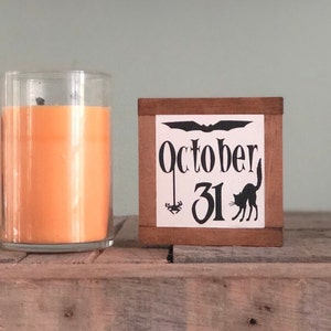 May include: A wooden block with a white background and black lettering that reads "October 31". A black bat and a black cat are also depicted on the block.