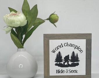 May include: A small wooden sign with a white background and black text. The sign says "World Champion Hide & Seek" and features a silhouette of a Bigfoot walking through a forest.