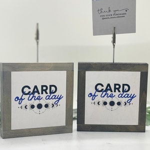 May include: Two wooden signs with a gray wash finish and a white background. The signs have a black frame and the text "CARD of the day" in black and blue lettering. The signs have a moon and star design.