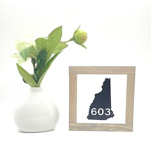 May include: A small, square wooden sign with a white background features a black silhouette of the state of New Hampshire and the number 603. A white vase holds a faux flower arrangement with green leaves and white flowers.