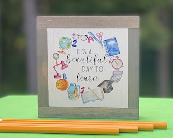May include: A wooden block with a watercolor illustration of school supplies and the text "It's a beautiful day to learn." The illustration includes a globe, glasses, a pen, scissors, a notebook, a clock, a calculator, a graduation cap, a book, a pencil, a backpack, and a basketball. Pencils are in the foreground.