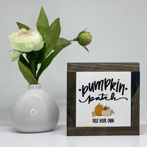 May include: A rustic wooden sign with a white background and black text that reads "pumpkin patch" with a handwritten font. The sign also features a small illustration of pumpkins and leaves. The text "Pick Your Own" is printed below the illustration.