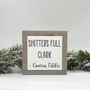 May include: A white wooden sign with the text "Shitters Full Clark - Cousin Eddie" in black lettering.