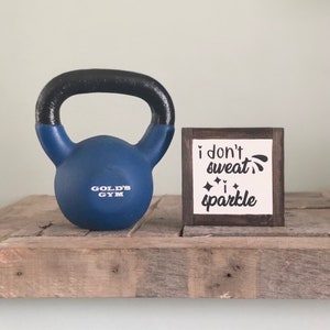 May include: A blue kettlebell with the words "Gold's Gym" printed on it sits on a wooden shelf next to a small wooden sign with the text "i don't sweat i sparkle".
