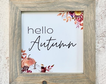 May include: Square wooden framed sign with the words "hello Autumn" in black script. The sign features watercolor pumpkins and floral accents in shades of orange, white, and burgundy. The frame is a light gray color.