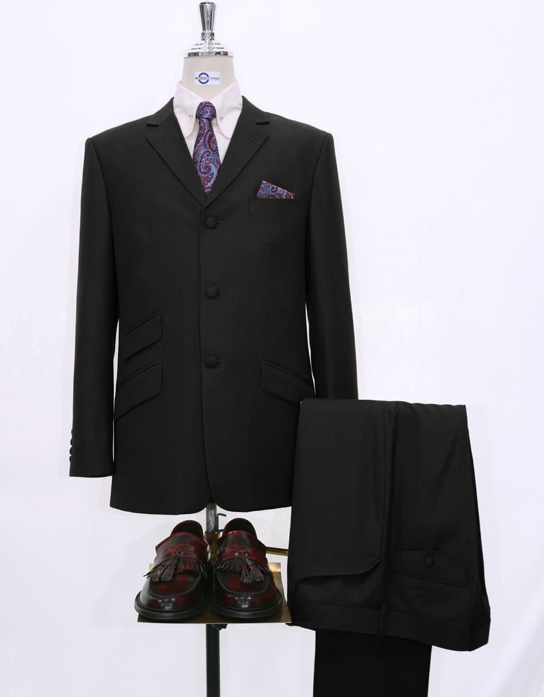 May include: A black suit with a three-button closure, a patterned tie, and a pocket square. The suit includes a jacket and matching trousers. The suit is displayed on a mannequin with a white shirt and brown tassel loafers.