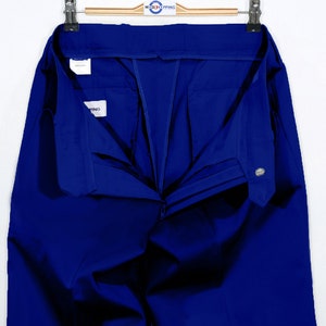 May include: A pair of blue pants with a zipper and button closure. The pants are folded in half and hanging on a wooden hanger.
