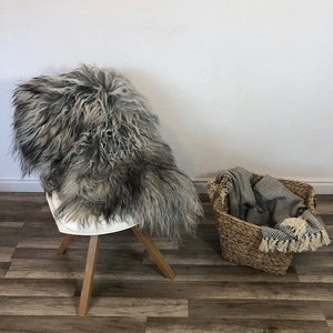 May include: A gray and black sheepskin rug draped over a white chair with wooden legs. A woven basket holds a folded blanket with fringe. The text "ExclusiveSheepskin" is at the bottom.