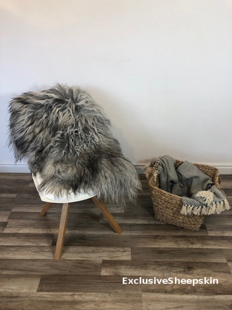 May include: A gray and black sheepskin rug draped over a white chair with wooden legs. A woven basket holds a folded blanket with fringe. The text "ExclusiveSheepskin" is at the bottom.
