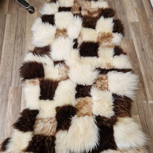 Pode incluir: Um tapete de pele de ovelha em patchwork em tons de castanho, creme e bege. O tapete é feito de pedaços retangulares e quadrados de pele, criando um padrão geométrico texturizado. O tapete está colocado num chão de madeira.