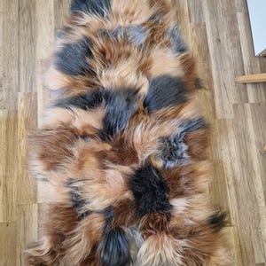May include: A rectangular sheepskin rug with a patchwork design. The rug features a mix of colors, including shades of brown, black, and tan. The fur is long and shaggy, creating a textured appearance. The rug is designed to be placed on the floor.