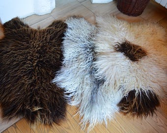 May include: Three sheepskin rugs in different colors: brown, white, and gray. The rugs are laid out on a wooden floor.