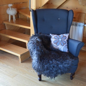 Op de afbeelding: Een donkerblauwe fauteuil met een hoge rugleuning en knoopbekleding. De stoel is bedekt met een pluche grijze deken en een kussen met bloemenprint. De stoel staat in een kamer met houten vloer en een houten trap op de achtergrond.