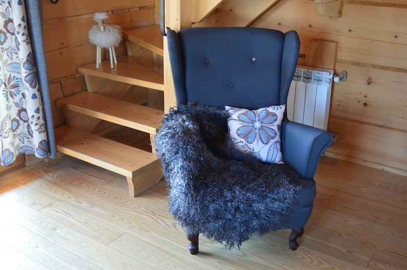 Op de afbeelding: Een donkerblauwe fauteuil met een hoge rugleuning en knoopbekleding. De stoel is bedekt met een pluche grijze deken en een kussen met bloemenprint. De stoel staat in een kamer met houten vloer en een houten trap op de achtergrond.