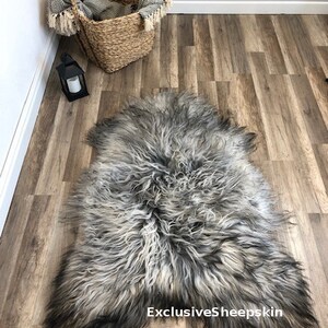 May include: A gray and black sheepskin rug with long, shaggy fur. The rug is displayed on a wood-look floor. A woven basket with a blanket and a black lantern are in the background. The text "ExclusiveSheepskin" is visible on the rug.