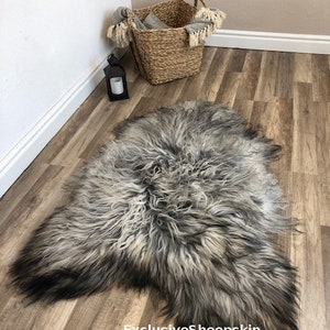 May include: A gray and black sheepskin rug lies on a wooden floor. The rug has long, shaggy fur with a gradient effect, transitioning from light gray to dark gray and black. A woven basket and a black lantern are in the background.