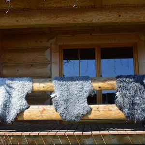 Op de afbeelding: Drie grijze schapenvachten hangen aan een houten leuning buiten een blokhut. De vachten hangen aan een houten leuning buiten een blokhut.