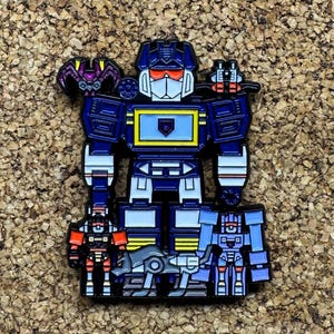 Puede incluir: Un pin de esmalte metálico que muestra un robot grande de color azul con un panel pectoral amarillo, visera naranja y cara blanca. El robot está rodeado por cuatro figuras robóticas más pequeñas en rojo, gris y azul, con una criatura morada.