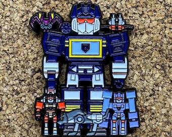May include: A metal enamel pin featuring a large blue robot character with a yellow chest panel, orange visor, and white face. The robot is surrounded by four smaller robot figures in red, grey, and blue, with a purple bat-like creature on the shoulder.