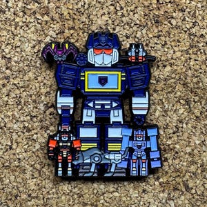 May include: A blue, white, and red enamel pin featuring a robot character with a rectangular chest and multiple smaller robot figures. The pin has a gold border and is set against a textured, brown background. The robot has a red visor.