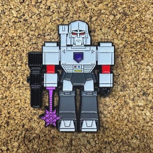 May include: A silver, gray, and black Megatron pin with red accents. The pin features a purple emblem on the chest and a purple weapon. The pin is a stylized representation of the Transformers character.