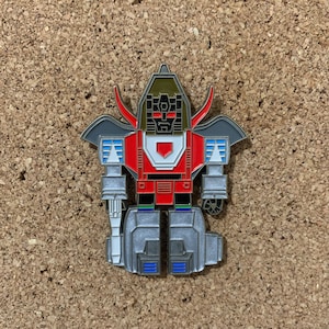 May include: A silver, red, and gold robot pin with a black face and a red and white chest plate. The robot has a cape and a sword.