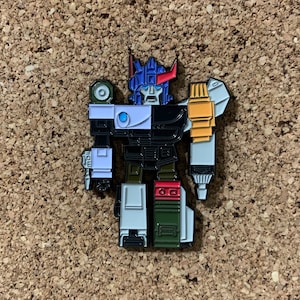 May include: A black, white, blue, red, and yellow enamel pin of a robot in a standing pose. The robot has a large head with a blue helmet and a red visor. The robot's arms are extended to the sides, and its legs are together.