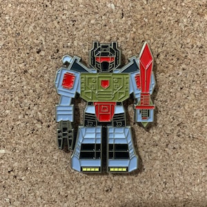 May include: A silver, black, red, and green robot pin with a red sword. The robot has a black head with a red visor and a green chest.