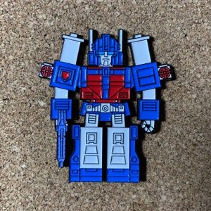 May include: A blue and red robot pin with a white face and a gun. The robot has a red chest and a blue body. The pin is made of metal and has a shiny finish.