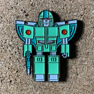 May include: A green and blue robot pin with red accents. The robot has a large head with a visor and a large body with wings. The robot is holding a gun in its right hand.