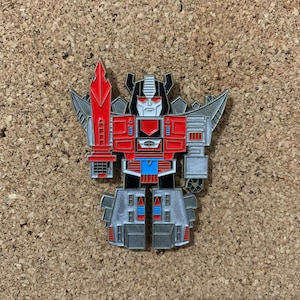 May include: A red and silver robot pin with a sword. The robot has a black face with white eyes and a red mouth. The robot is standing on a silver base.