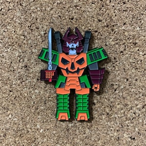 May include: A green, orange, and black enamel pin of a robot with a pumpkin head. The robot is holding a sword and has a samurai-like design.