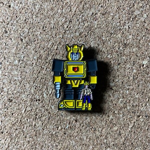 May include: A yellow and black robot pin with a smaller yellow and black robot figure standing in front of it. The larger robot has a drill on its arm and the smaller robot has a tool belt.