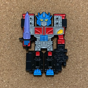 May include: A blue, red, and gray robot pin with a sword. The robot has a yellow chest plate and a blue head with a red visor.