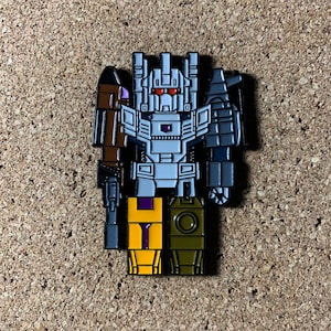 May include: A gray, white, purple, yellow, and brown robot pin with a red eye. The robot is holding a gun in its right hand and a weapon in its left hand.