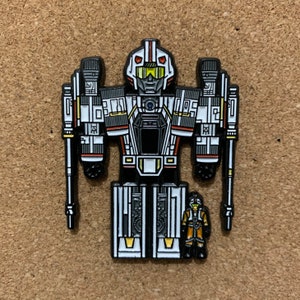 May include: A black, white, and red robot pin with a yellow visor and a smaller pilot figure. The robot is holding two long, black weapons.