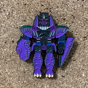 May include: A purple and black robot pin with a dinosaur head on its left arm and a shield on its right arm. The robot has red eyes and yellow claws. The pin is on a brown, textured surface.