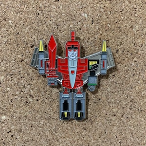 May include: A red and silver robot pin with a sword and wings. The robot has a yellow face and a red chest. The pin is a collectible item.