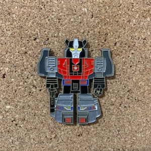 May include: A red, black, and silver robot pin with a yellow visor. The robot has a black face with a white mouth and a black cross on its chest.
