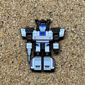 May include: A white and black robot pin with a blue visor and red chest emblem. The robot is in a standing pose with a gun in its right hand.